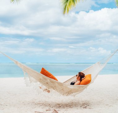 Mauritius sahilindeki kumlu beyaz plajda hasır hamakta dinlenen ve geniş okyanus manzaralı dalgaların tadını çıkaran genç bir kadın. Egzotik ülkeler tatil ve ruh sağlığı konsepti imajı.
