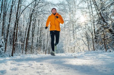Orta yaşlı, gülümseyen koşucu parlak turuncu rüzgarsız ceket giymiş dayanıklı güneşli ve karlı bir günde karlı ormanda hızla koşuyor. Sportif aktif insanlar ve kış eğitim imajı.