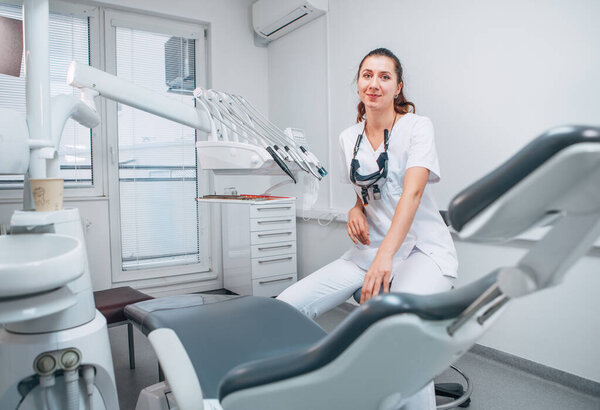 Portraait of sincerely smiling young dentist woman dressed white medical scrubs uniform sitting in modern dental clinic next to stomatology  chair. Health care and medicare industry concept image.
