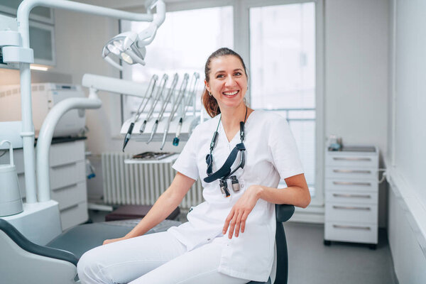 Portraait of sincerely smiling young dentist woman dressed white medical scrubs uniform sitting in modern dental clinic next to stomatology  chair. Health care and medicare industry concept image.