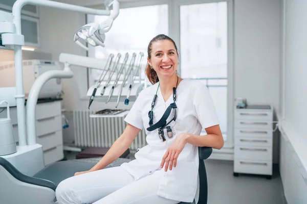 心から笑顔の若い歯科医師の女性のポートレイトは 現代の歯科診療所に座っている白い医療スクラブのユニフォームを着て ストーマトロジーチェアの隣にあります メディケア業界のコンセプトイメージ ストックフォト