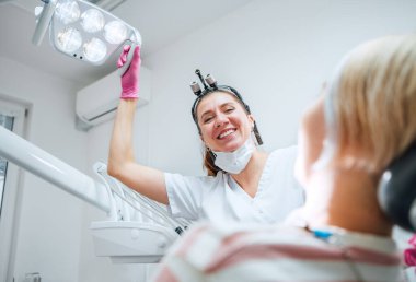 Modern tıp koğuşunda dişçi muayenehanesinde hasta randevusu. Dişçi doktor içtenlikle gülümsüyor, diş ameliyatına hazır genç bir kadına diş ışığını doğrultuyor. Sağlık ve sağlık sektörü imajı.