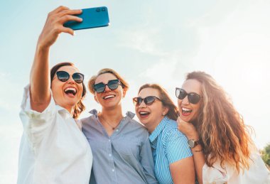 Güneş gözlüklü, gülümseyen dört kadının portresi. Birbirlerine sarılıyorlar ve doğa yürüyüşü sırasında modern akıllı telefon kullanarak selfie çekiyorlar. Kadınların dostluğu, ilişkileri ve mutluluk konsepti imajı