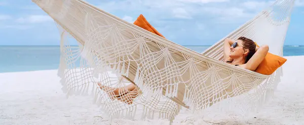 Young woman relaxing in wicker hammock on the sandy beach on Mauritius coast and enjoying wide ocean view waves. Exotic countries vacation and mental health concept image.