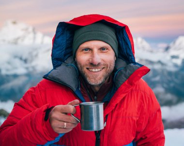 Yüksek irtifada kameraya gülümseyen dağcı portresi. Kırmızı, ılık şafak ceketi giymiş. Elinde dağ manzaralı bir fincan sıcak çay tutuyor. Mera Tepesi Kampı 5700 metre, Himalayalar, Nepal.