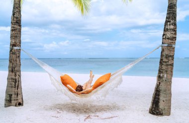 Mauritius sahilindeki kumlu beyaz plajda hasır hamakta dinlenen ve geniş okyanus manzaralı dalgaların tadını çıkaran genç bir kadın. Egzotik ülkeler tatil ve ruh sağlığı konsepti imajı.