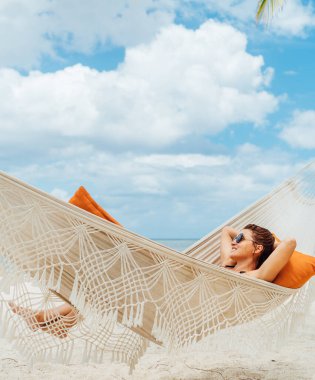 Young woman relaxing in wicker hammock on the sandy beach on Mauritius coast and enjoying wide ocean view waves. Exotic countries vacation and mental health concept image.