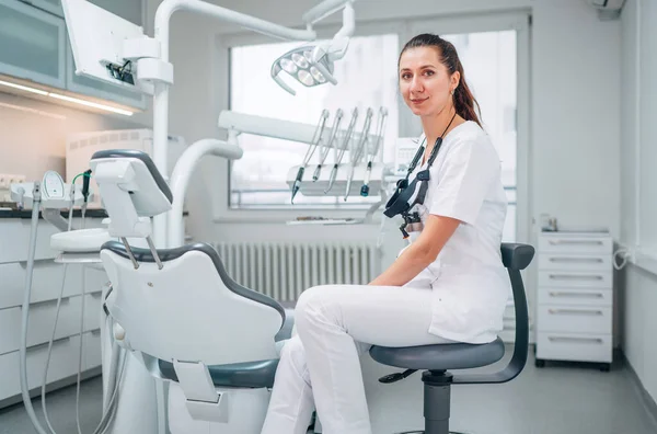 心から笑顔の若い歯科医師の女性のポートレイトは 現代の歯科診療所に座っている白い医療スクラブのユニフォームを着て ストーマトロジーチェアの隣にあります メディケア業界のコンセプトイメージ ストック画像