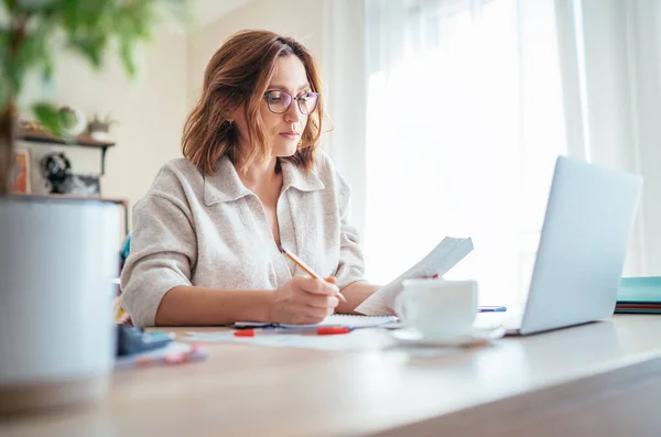 Beautiful middle-aged woman in glasses counting family business budget gazing at monthly bills and documents at home living room office. Small business home finances, money savings concept image