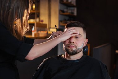 Uzun saçlı genç bayan berber sandalyedeyken sakallı adamın yüzünü nazikçe tıraş ediyor. Modern hafif siyah tarzı berber dükkanı iç mimarisi. Saç ve cilt bakımı, yerel küçük işletme konsepti..