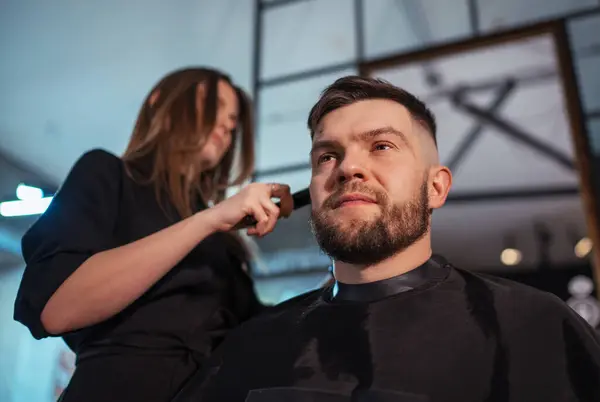 Stern bearded Man portrait while undercut hairstyle hairdressing in Hair Salon by young hairdresser female. Modern low light black style barber shop interior. Haircare service small business concept