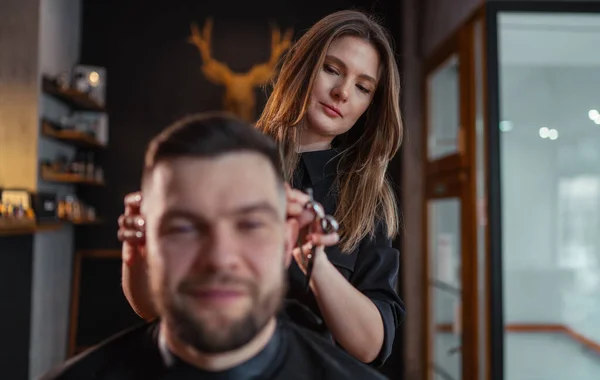Young female barber portrait she gently making undercut hairstyle for bearded man in Hair Salon Chair. Modern low light black style barber shop interior. Haircare service local small business concept