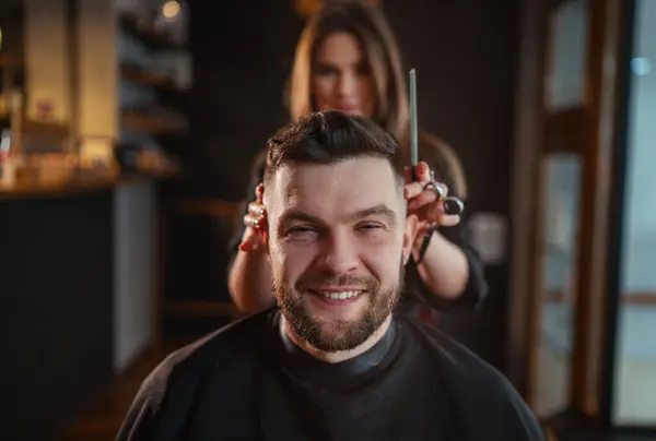 Smiling bearded Man portrait while undercut hairstyle hairdressing in Hair Salon by young hairdresser female. Modern low light black style barber shop interior. Haircare service small business concept