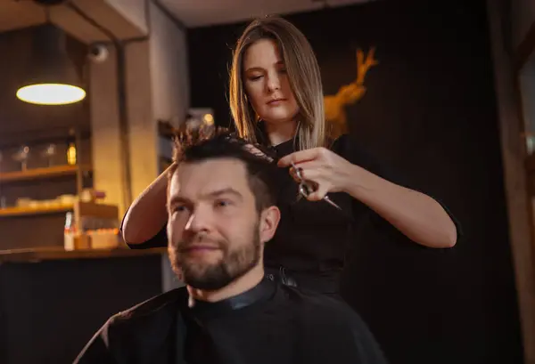 Young long haired female barber gently makes undercut hairstyle using scissors and comb bearded happy man in Salon Chair. Modern black-style barber shop. Hair Care service local small business concept
