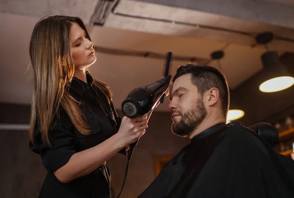 Young long long-haired female hairdresser hairstyling drying hair with professional hair dryer bearded man in Salon Chair. Modern black-style barber shop.Hair Care service local small business concept
