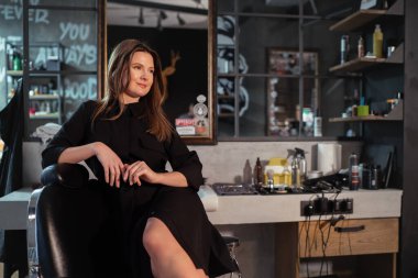 Güzel, uzun saçlı, yumuşak gülümseyen, siyah elbiseli bir kuaför kadının portresi. Berber koltuğunda oturmuş, sıcak ışıkla dinleniyor. Saç bakımı, kadın güzelliği, moda konsepti.