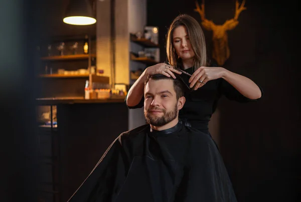 Young long haired female barber gently makes undercut hairstyle using scissors and comb bearded happy man in Salon Chair. Modern black-style barber shop. Hair Care service local small business concept