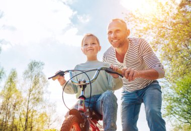 Gülümseyen baba kızına bisiklet sürmeyi öğretiyor. Onlar yaz şehir parkında birliktelikten hoşlanırlar. Mutlu çocukluk konsepti düşük açı görüntü.