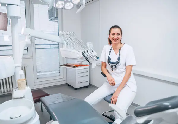 心から笑顔の若い歯科医師の女性のポートレイトは 現代の歯科診療所に座っている白い医療スクラブのユニフォームを着て ストーマトロジーチェアの隣にあります メディケア業界のコンセプトイメージ ロイヤリティフリーのストック画像