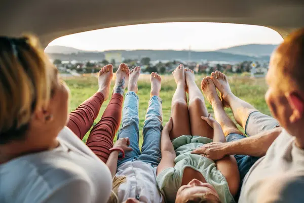 Four-person family lying in the car trunk and fooling around raising feet up. The young caucasian couple has an auto trip break. Family values, traveling, and friendship concept.