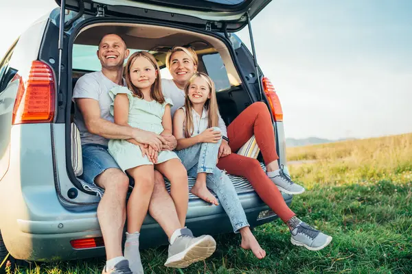 自動車トロフィーの間に車のトランクの中に座っている小さな娘たちと幸せな若いカップルの肖像画 笑ったり 笑ったり しゃべったりしている 家族の価値観 旅行コンセプト — ストック写真