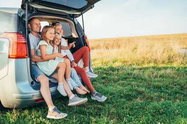 Portrait of Happy young couple with little daughters sitting inside car trunk during auto trop. They are smiling, laughing and chatting. Family values, traveling concept.