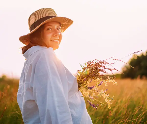 Sincerely smiling young pregnant woman dressed in light summer clothes and straw hat walking by high green grass meadow with wildflowers bouquet in evening sunset hours. Human in nature concept image.