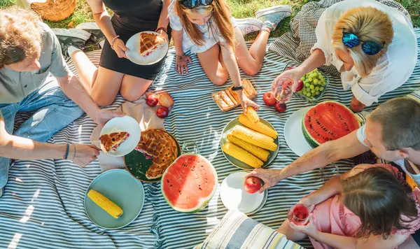 Top view of big family sitting on the picnic blanket in city park. They are eating boiled corn, apples, peaches, pastries and watermelon. Family values and outdoor activities concept