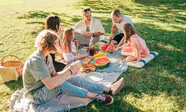 Big family sitting on the picnic blanket in city park during weekend Sunday sunny day. They are smiling, laughing and eating boiled corn and watermelon. Family values and outdoors activities concept.