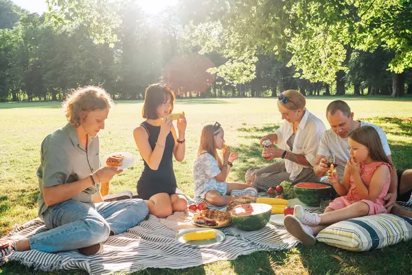 日曜日の晴れた日には 市内の公園のピクニックブランケットに座っている大きな家族 彼らは笑顔で 笑って ゆでたトウモロコシとスイカを食べている 家族の価値観とアウトドア活動のコンセプト ストック画像