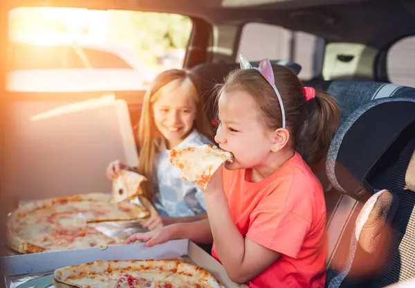 Portrait of two positive smiling sisters eating just cooked italian pizza sitting in child car seats on car back seat. Happy childhood, fastfood eating or auto jorney lunch break concept image.