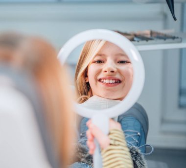 Stomatoloji kliniği koltuğunda oturan küçük kız dişlerini gösterdikten sonra aynaya bakıp gülümsüyor. Sağlık, çocuk sağlığı ve sağlık sektörü konsepti
