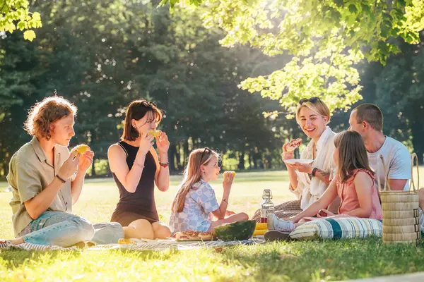日曜日の晴れた日には 市内の公園のピクニックブランケットに座っている大きな家族 彼らは笑顔で 笑って ゆでたトウモロコシとスイカを食べている 家族の価値観とアウトドア活動のコンセプト ストック写真