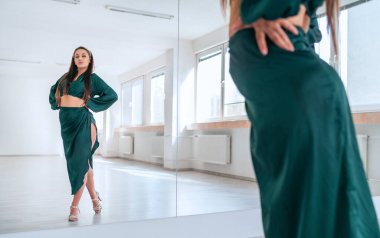 Koyu yeşil Latin dans elbisesi giymiş zarif bir kadın büyük ayna duvarlı zarif dans sırasında aynaya poz veriyor. Dans sırasında insanların ifadeleri, kadın vücudunun konsept imajının güzelliği.