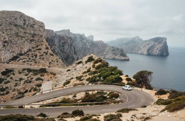 Serpentine kıvrımlı asfalt dağ yolunun yanından geçen yalnız, küçük, beyaz bir araba. Kayalık sahili olan deniz manzaralı deniz feneri yakınlarında. Mallorca Adası, Balear Adaları, İspanya.