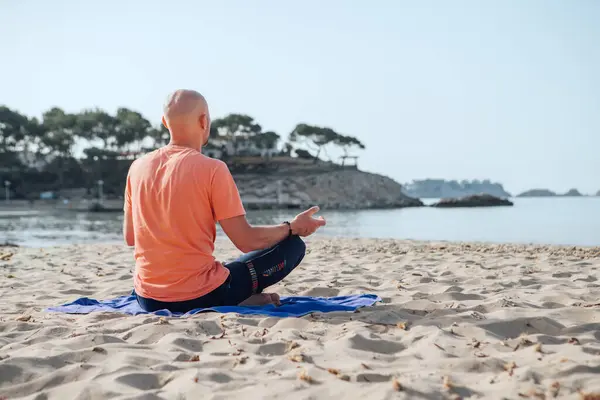 Calm man sitting in lotus position, doing deep breathing exercises and meditating in early morning hours on the sandy beach with calm sea waves. Mental health, people relaxing, traveling concept.