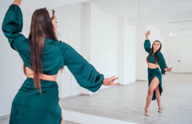Koyu yeşil Latin dansı elbisesi giymiş zarif bir kadın büyük ayna duvarlı beyaz renkli salonda zarif danslar yapıyor. Dans sırasında insanların ifadeleri, kadın vücudunun konsept imajının güzelliği.