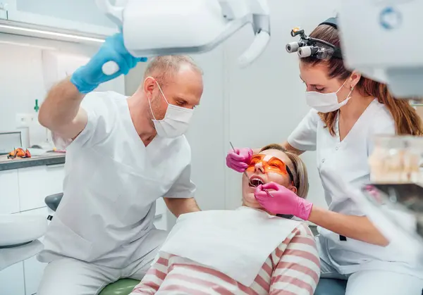 牙科诊所病人预约 戴放大镜的牙医妇女使用医疗工具进行牙齿预防 年轻人的助手指着灯 医疗保健和医疗行业概念形象 — 图库照片