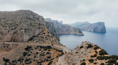 Yalnız beyaz araba serpentin kıvrımlı asfalt dağ yolunda Cap de Formentor kayalık sahilinin deniz feneri yakınında. Mallorca Adası, Balear Adaları, İspanya. Uçan İHA hava aracı görüntüsü.