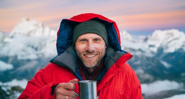 Yüksek irtifada kameraya gülümseyen dağcı portresi. Kırmızı, ılık şafak ceketi giymiş. Elinde dağ manzaralı bir fincan sıcak çay tutuyor. Mera Tepesi Kampı 5700 metre, Himalayalar, Nepal.