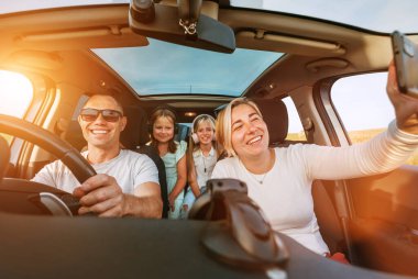 Otomobil kazasında iki kızı olan mutlu genç bir çift. Gülümsüyorlar, gülüyorlar ve akıllı telefon kullanarak selfie çekiyorlar. Aile değerleri, seyahat, sosyal medya ve yeni teknoloji kavramları.