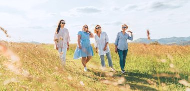 Dört neşeli kadının portresi el ele tutuşup yüksek yeşil bir çayırda yürüyor. Kameraya bakıyorlar. Kadınların dostluğu, ilişkileri ve mutluluk konsepti imajı.