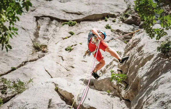 ベレー帽デバイスとクライミングハーネスロープを使用して崖の岩壁から身を隠しながら 保護ヘルメットのアクティブなクライマーの中年の男 アクティブな極端なスポーツの時間の支出の概念 ストック写真