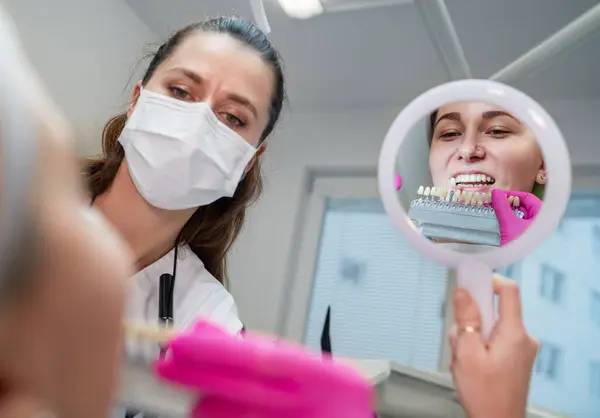 若い女性は歯科診療所の椅子に座り 鏡に微笑み 歯を示し 陰のタブで歯の色を選択しました ヘルスケア 口の健康 メディケア業界のコンセプトイメージ ストック画像