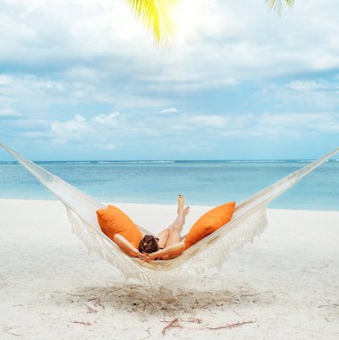 Mauritius sahilindeki kumlu beyaz plajda hasır hamakta dinlenen ve geniş okyanus manzaralı dalgaların tadını çıkaran genç bir kadın. Egzotik ülkeler tatil ve ruh sağlığı konsepti imajı.