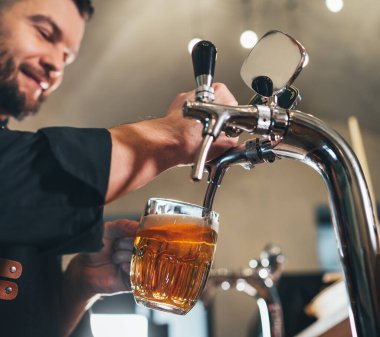 Gülümseyen şık sakallı barmen siyah üniformalı önlüklü barmen bardaki cam bardağa bira tıklatıyor. Başarılı insanlar, bira tüketimi, içecek endüstrisi imajı