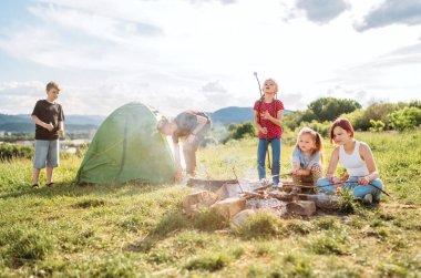 İki erkek kardeş yeşil çadırı kurarken, üç kız kardeş neşeyle gülüyor ve kamp ateşi üzerinde şekerleme ve şekerleme pişiriyor. Mutlu aile piknik kampı konsepti..