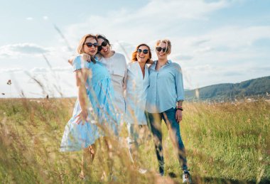 Dört neşeli kadının portresi el ele tutuşup yüksek yeşil bir çayırda yürüyor. Kameraya bakıyorlar. Kadınların dostluğu, ilişkileri ve mutluluk konsepti imajı.