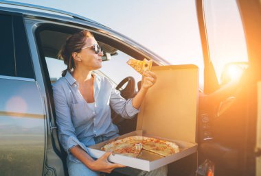 Orta yaşlı kadın yemek molasında sürücü koltuğuna oturmuş İtalyan pizzası pişiriyor ve günbatımının tadını çıkarıyor. Otomobil yolculuğu, fast food yeme ya da araba Jorney yemek molası konsepti.