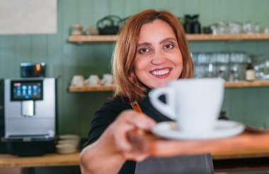 Cheerfully smiling barista female giving white cup aromatic espresso at the coffee shop counter. Happy people, coffee consumption, beverages industry and small business concept image. clipart